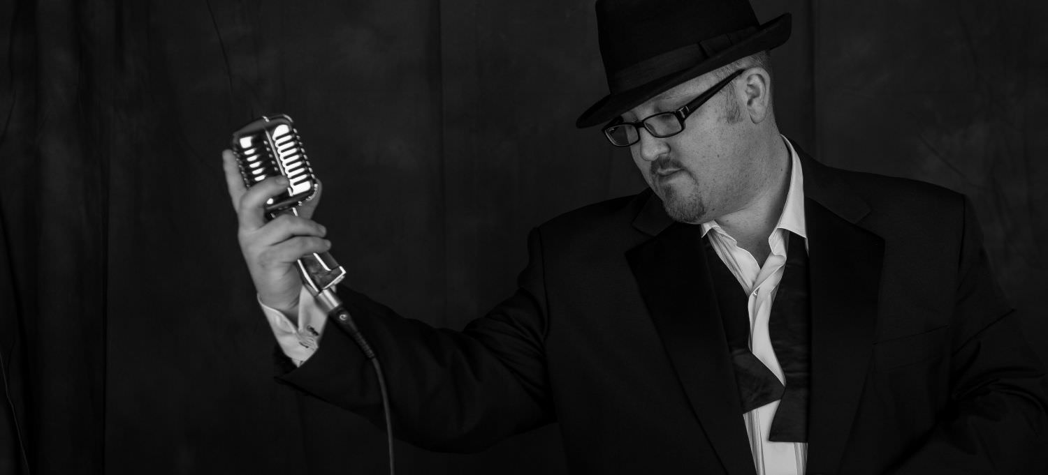 Jon paul swing singer in profile, wearing open bow tie under tuxedo while holding out vintage microphone near south wales.