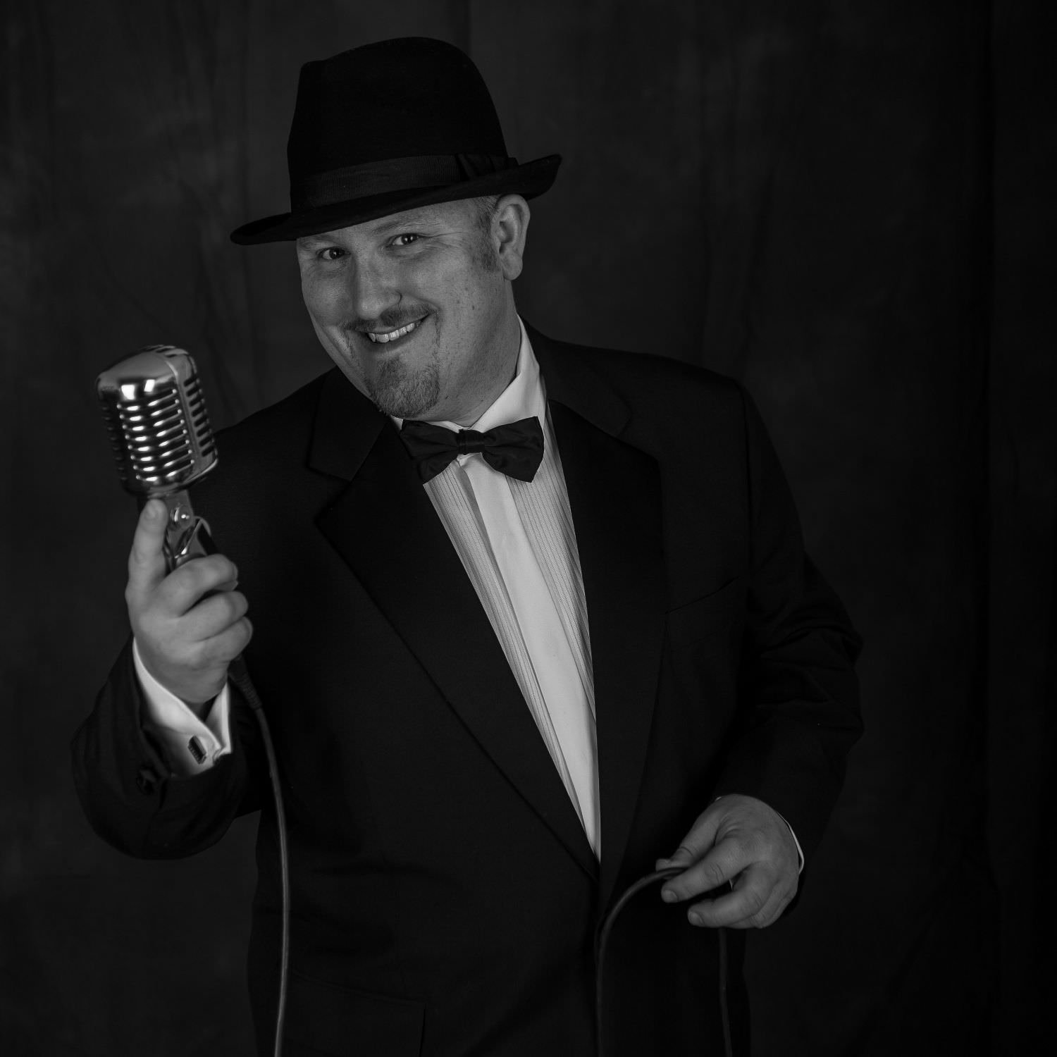 A smiling Jon paul rat pack singer in trilby hat and tuxedo with bow tie holds up 1950s microphone.