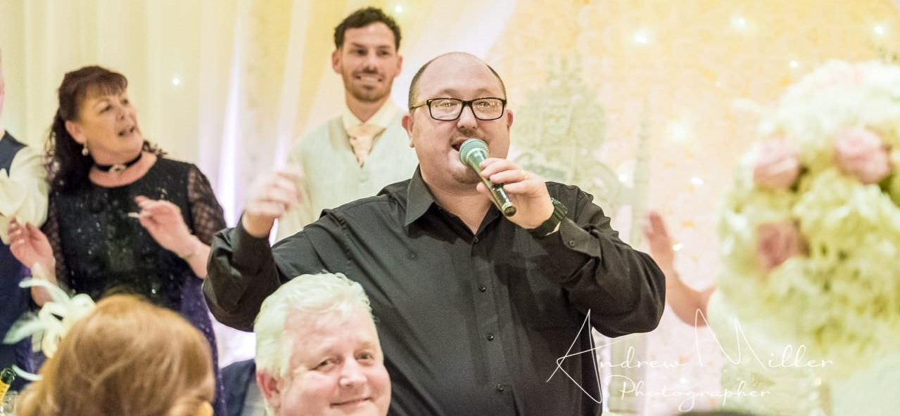 Groom and mother smile in background as jon paul in black shirt sings by wedding guests in front.