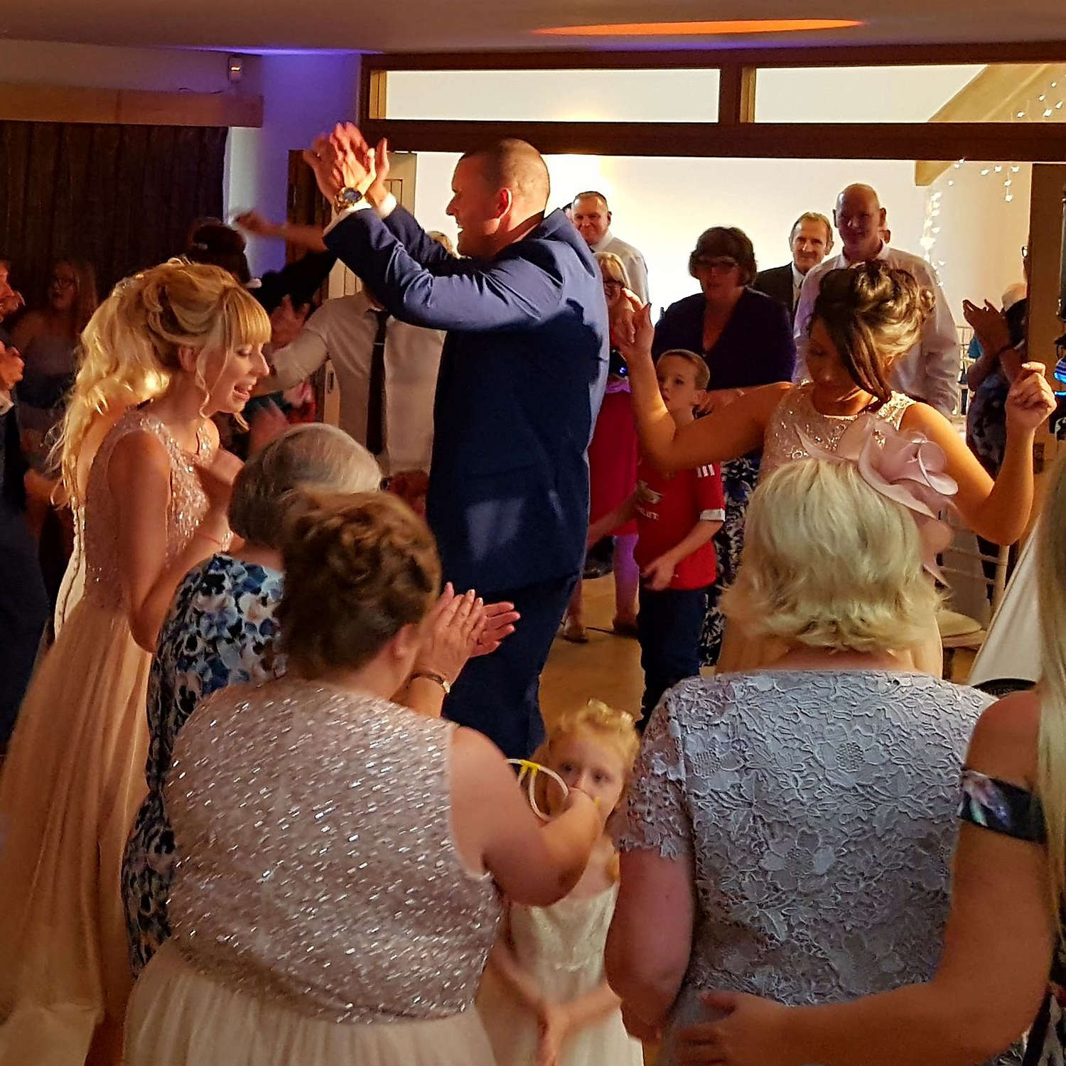 Happy dancing wedding guests surround smiling groom who claps his hands in the air at wedding near bristol.
