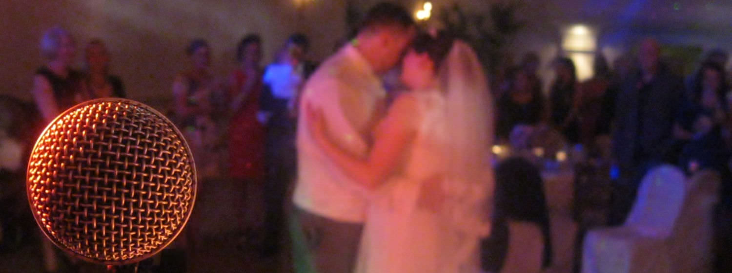 A microphone in the left foreground reveals bride and groom embraced in centre of dance floor with friends gathered behind.
